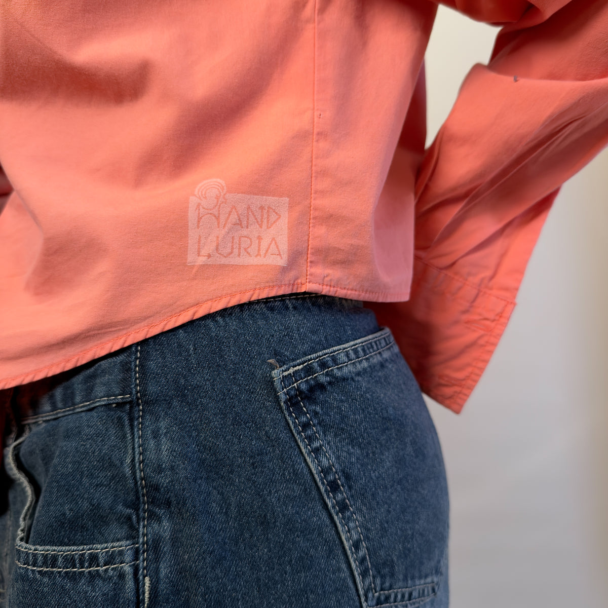 Detail view of Songbird Bloom by HANDLURIA, showing the coral shirt's curved hem and pale emblem.